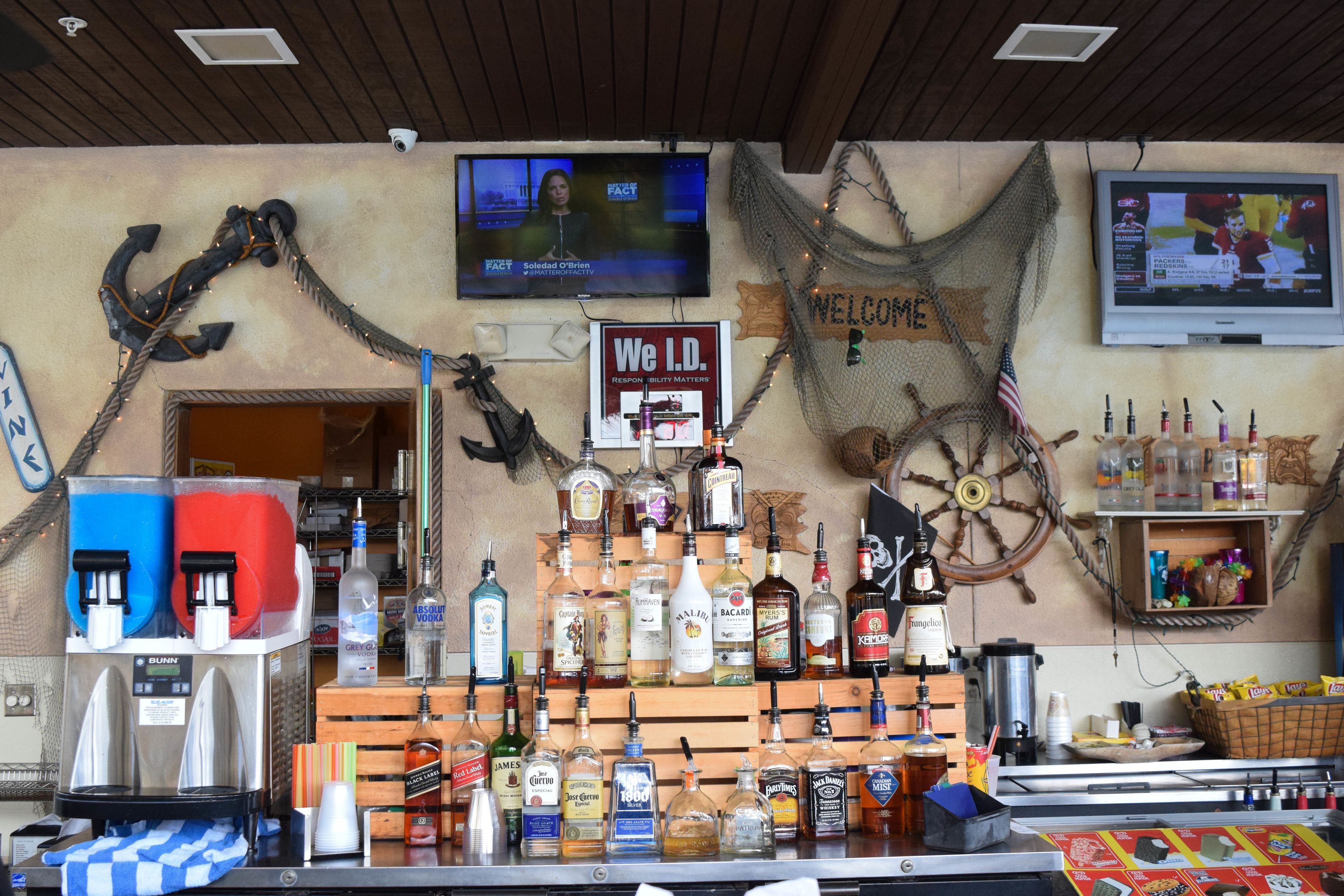 Eine Bar im maritimen Stil, mit Schiffsschwengel und Anker-Dekorationen. Verschiedene Flaschen und ein Fernsehbildschirm, der einen Nachrichtensender zeigt.