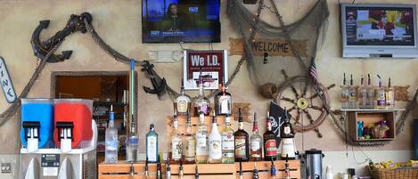 A bar area with a nautical theme, including a ship wheel and anchor decorations. There are various bottles and a television screen displaying a news channel.