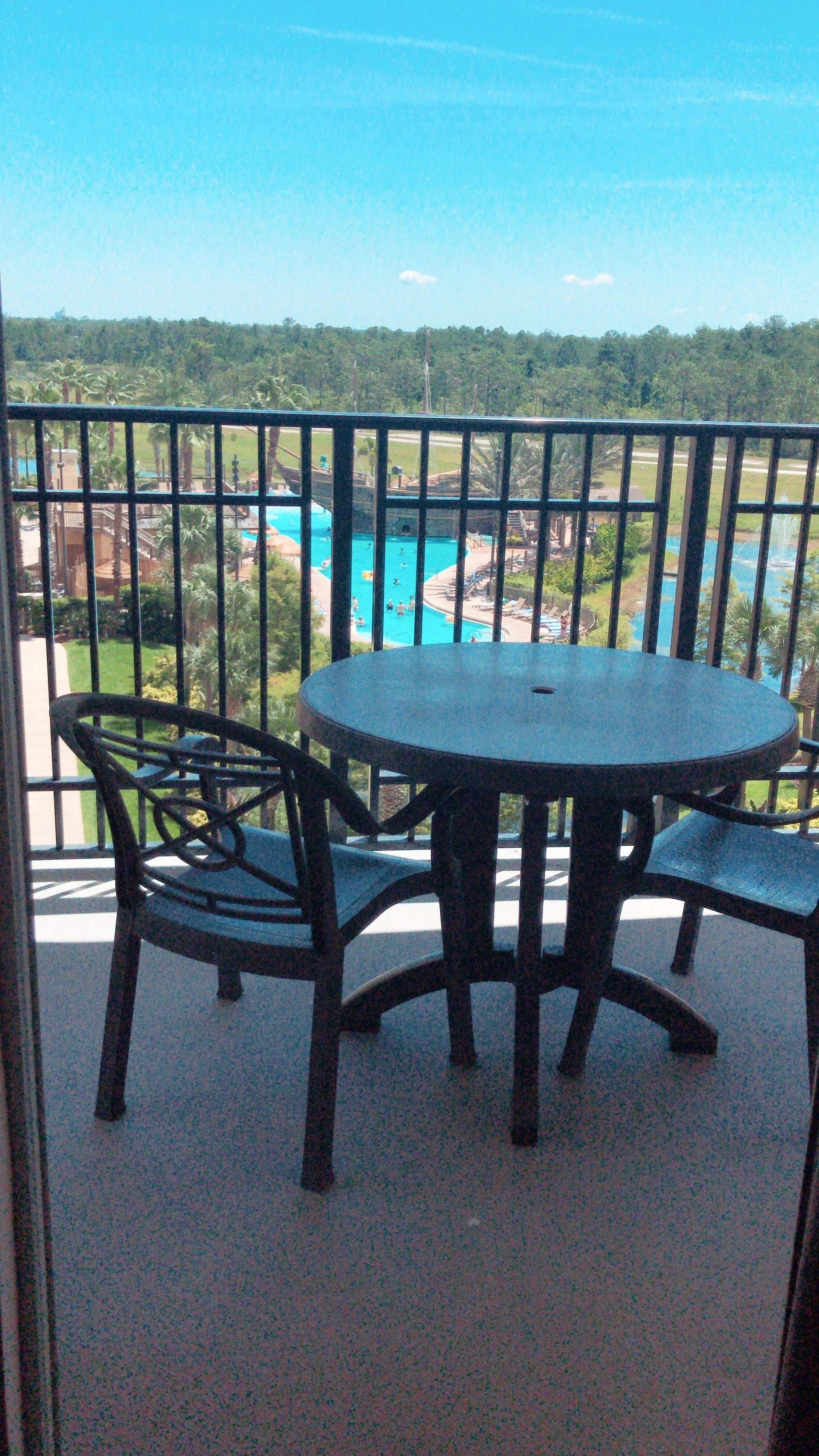 A balcony with a round table and two chairs, overlooking a swimming pool and lush greenery.