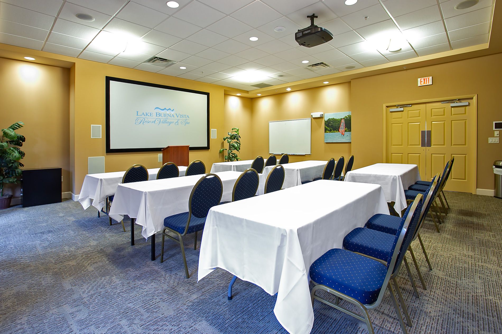 Une salle de conférence équipée de plusieurs tables et chaises, d’un écran de projection et d’un panneau indiquant la sortie.