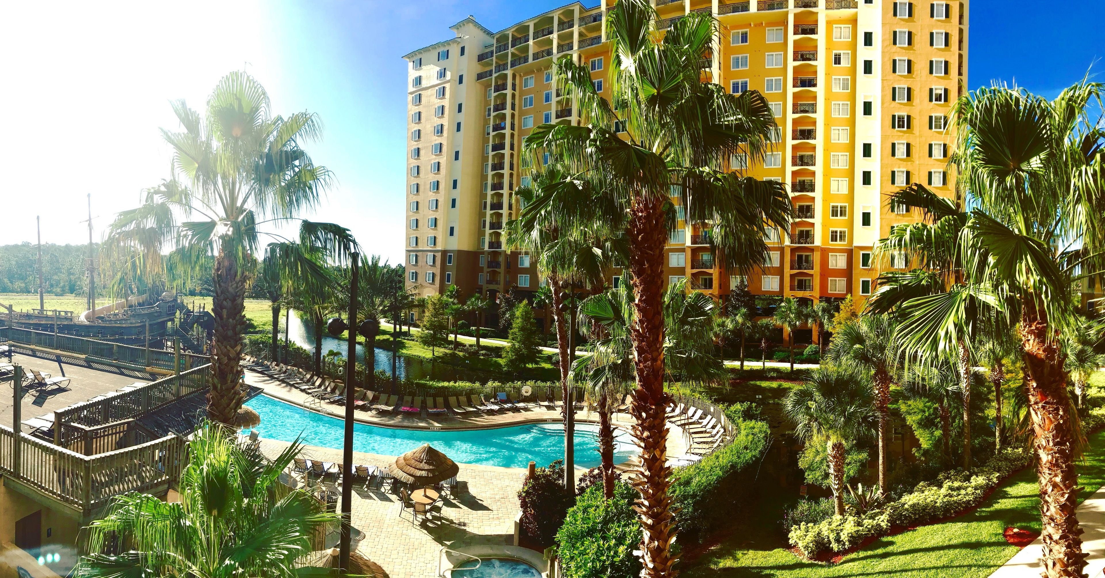 Un hotel con piscina, palmeras y un edificio grande al fondo.