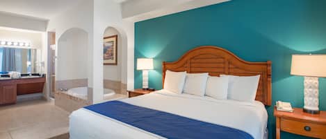 Habitación de hotel con una cama grande, cabecera de madera, dos lámparas de noche, mesitas de noche, baño con bañera y tocador con espejo e iluminación.
