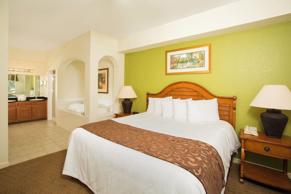 A hotel room with a large bed, a wooden headboard, two bedside lamps, a nightstand, a mirror, and a green accent wall.