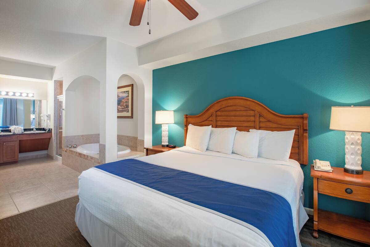 A hotel room with a large bed, a wooden headboard, a bathroom with a bathtub, and a vanity with a mirror.