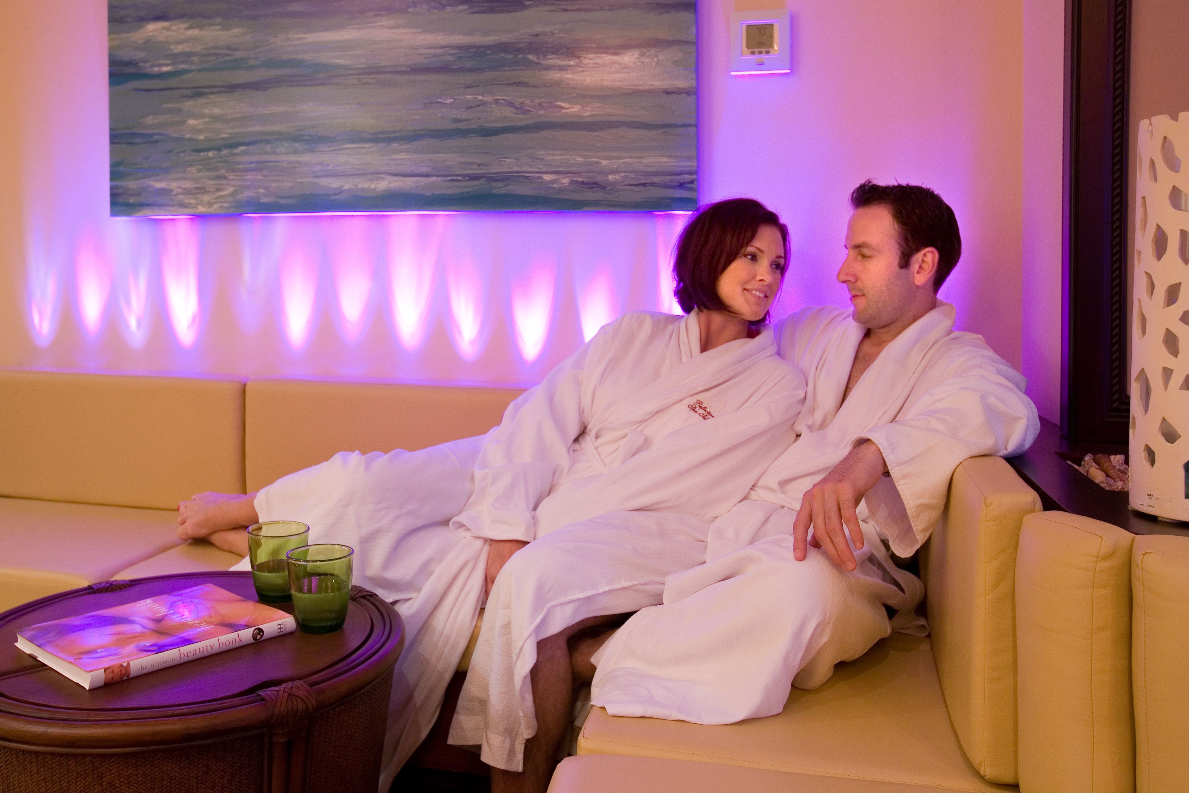 A couple in white robes sitting on a yellow couch in a room with a painting on the wall and a small table with a book and glasses.