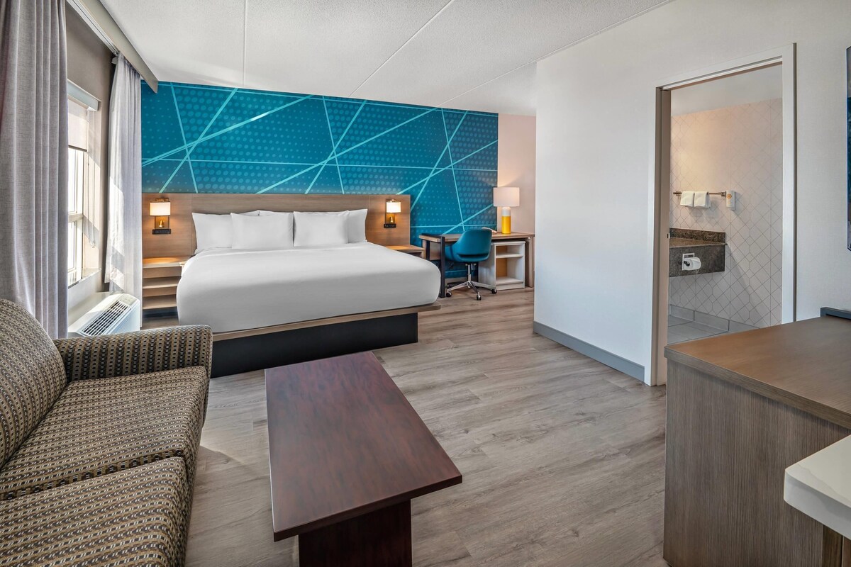 A hotel room with a bed, a desk, a chair, a sofa, and a bathroom visible through an open door.