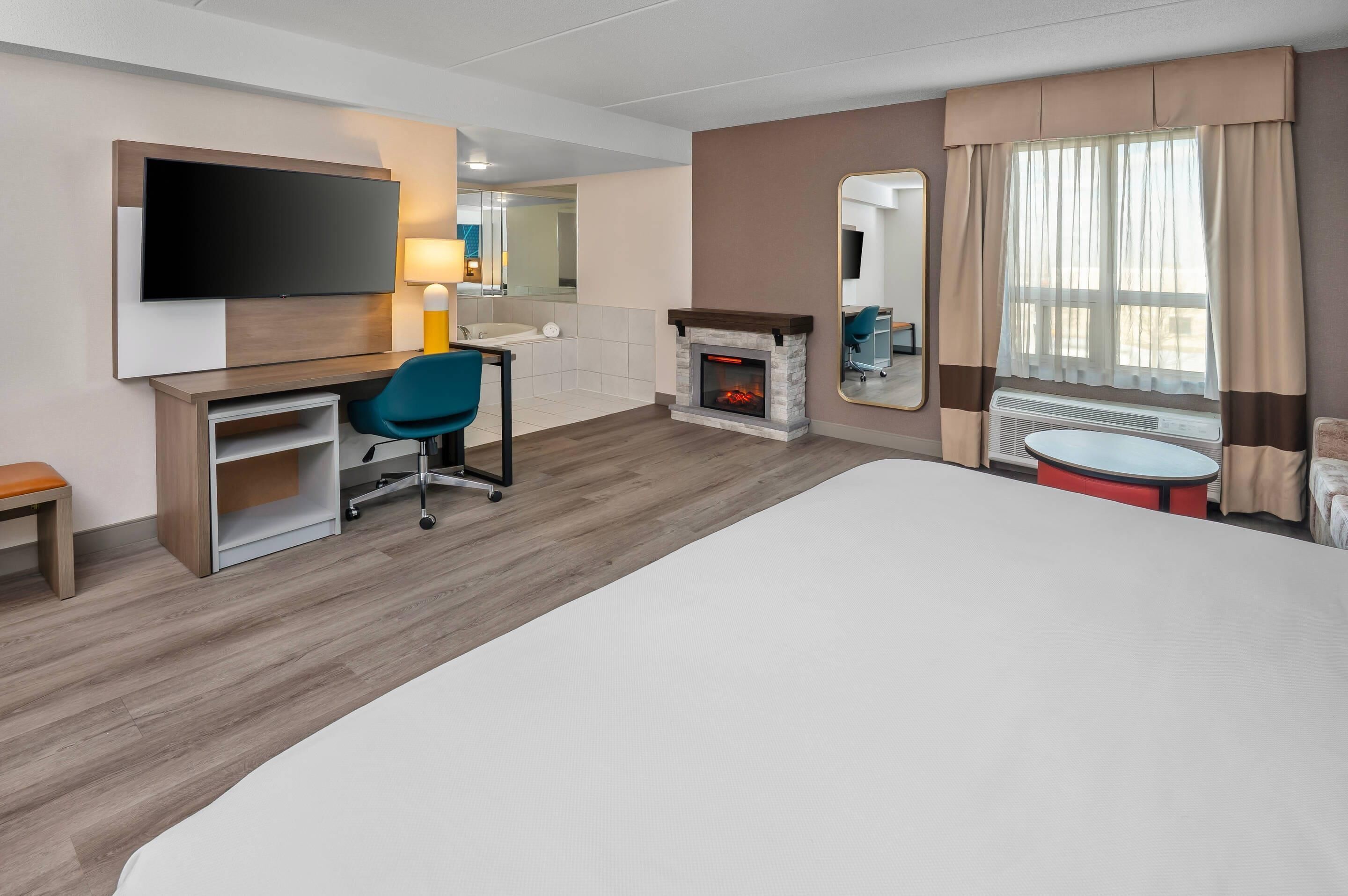 A hotel room with a large bed, a desk with a chair, a flat-screen TV, a fireplace, a large mirror, and a window with curtains.
