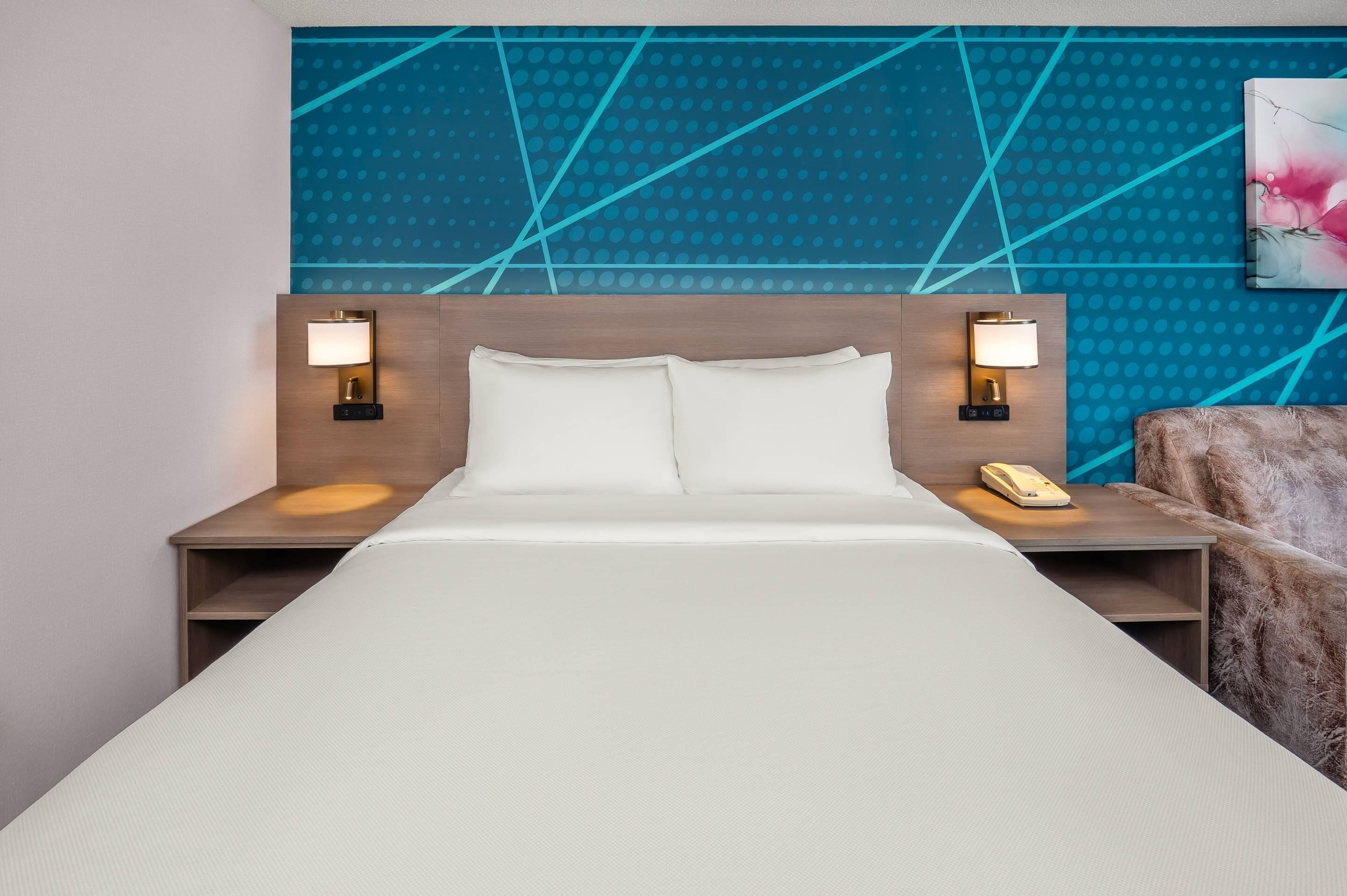 A hotel room with a bed, two bedside tables with lamps, a phone, and a wall with a geometric pattern.