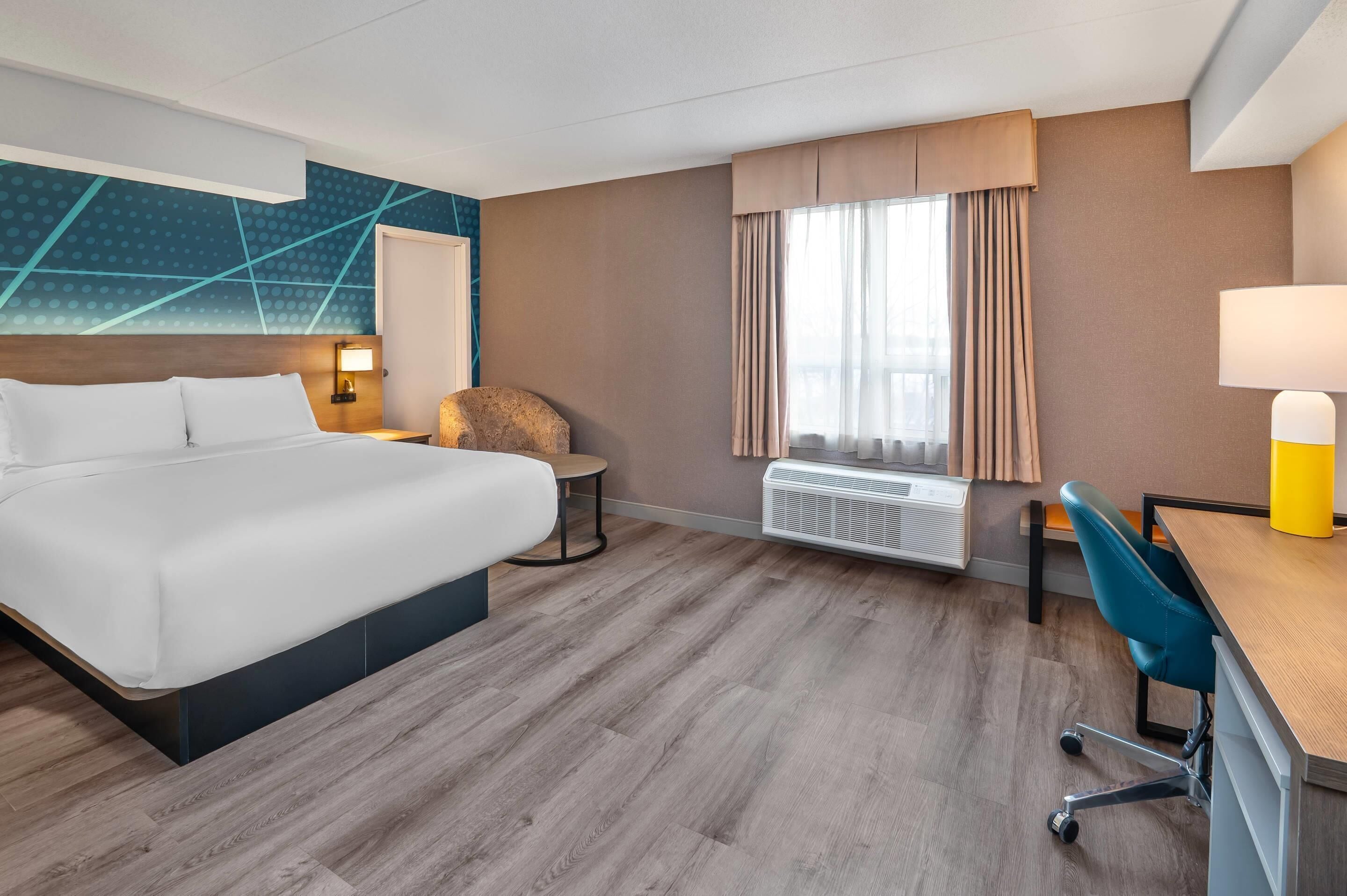 Una habitación de hotel con una cama grande, un escritorio, una silla, una mesa redonda, aire acondicionado instalado en la pared y una ventana con cortinas.