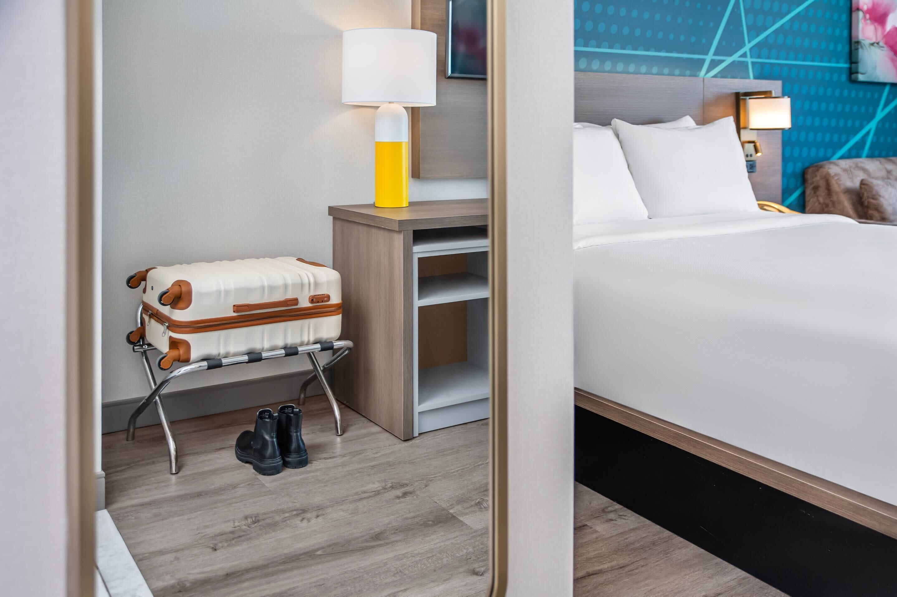 Hotellihuone, jossa on sänky, matkalaukku matkarullikossa, lamppu ja seinä, jossa on geometrinen kuvio.
