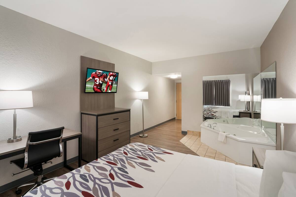 A modern hotel room with a flat-screen TV, a desk with a chair, a large bed with a patterned bedspread, a glass-enclosed jacuzzi, and a large mirror.