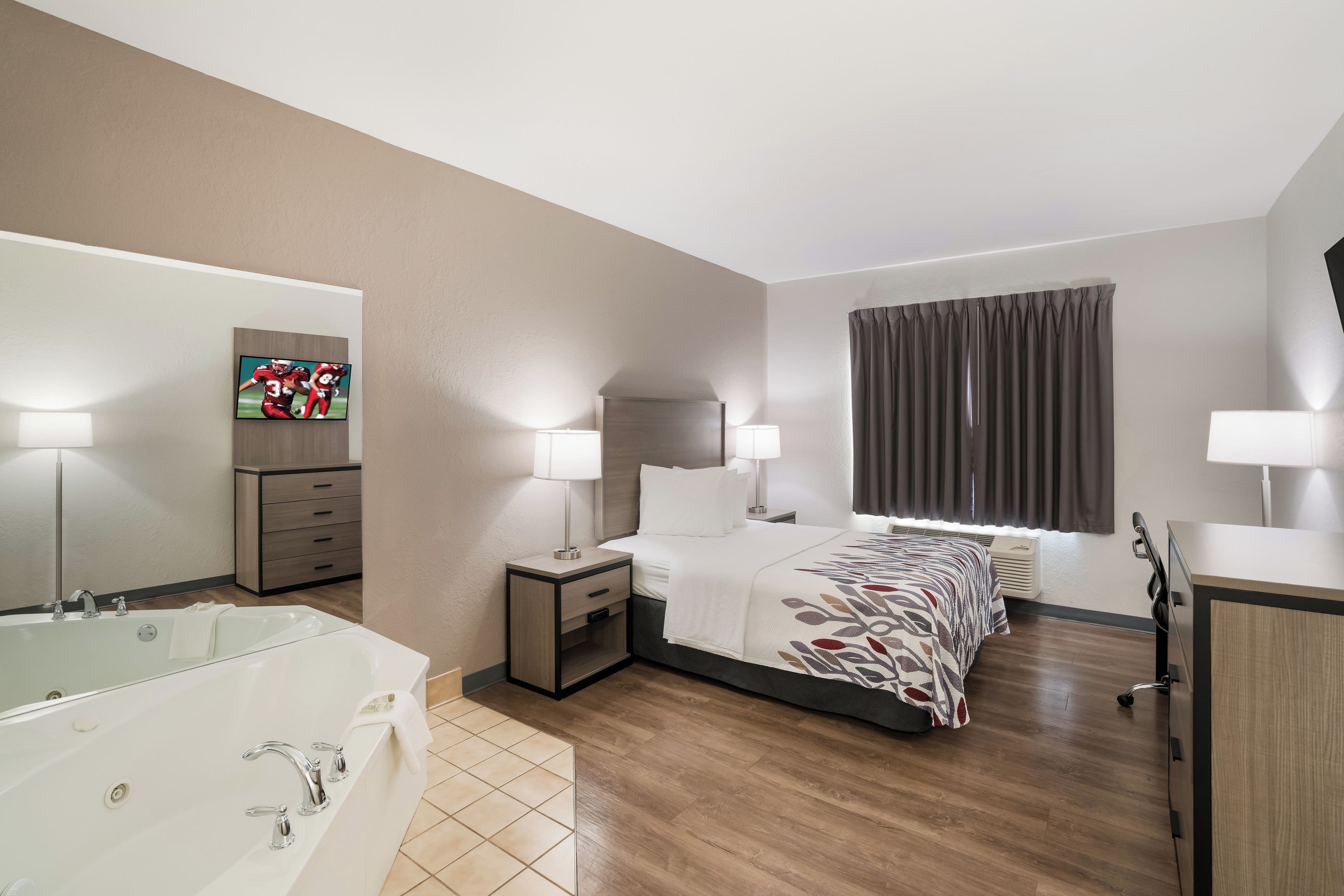 Een hotelkamer met een bed, nachtlampjes, een televisie, een badkamer met bad en een groot raam met gordijnen.