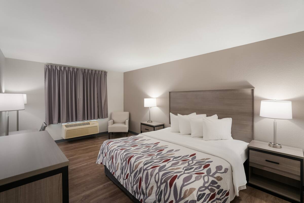 A hotel room with a bed, bedside tables, a desk, a chair, a window with curtains, and a wall-mounted air conditioner.