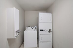 A laundry room with a stacked washer and dryer, a wall-mounted drying rack, and a door with a handle. - Red Roof Inn & Suites Carrollton, GA–West Georgia (Carrollton)