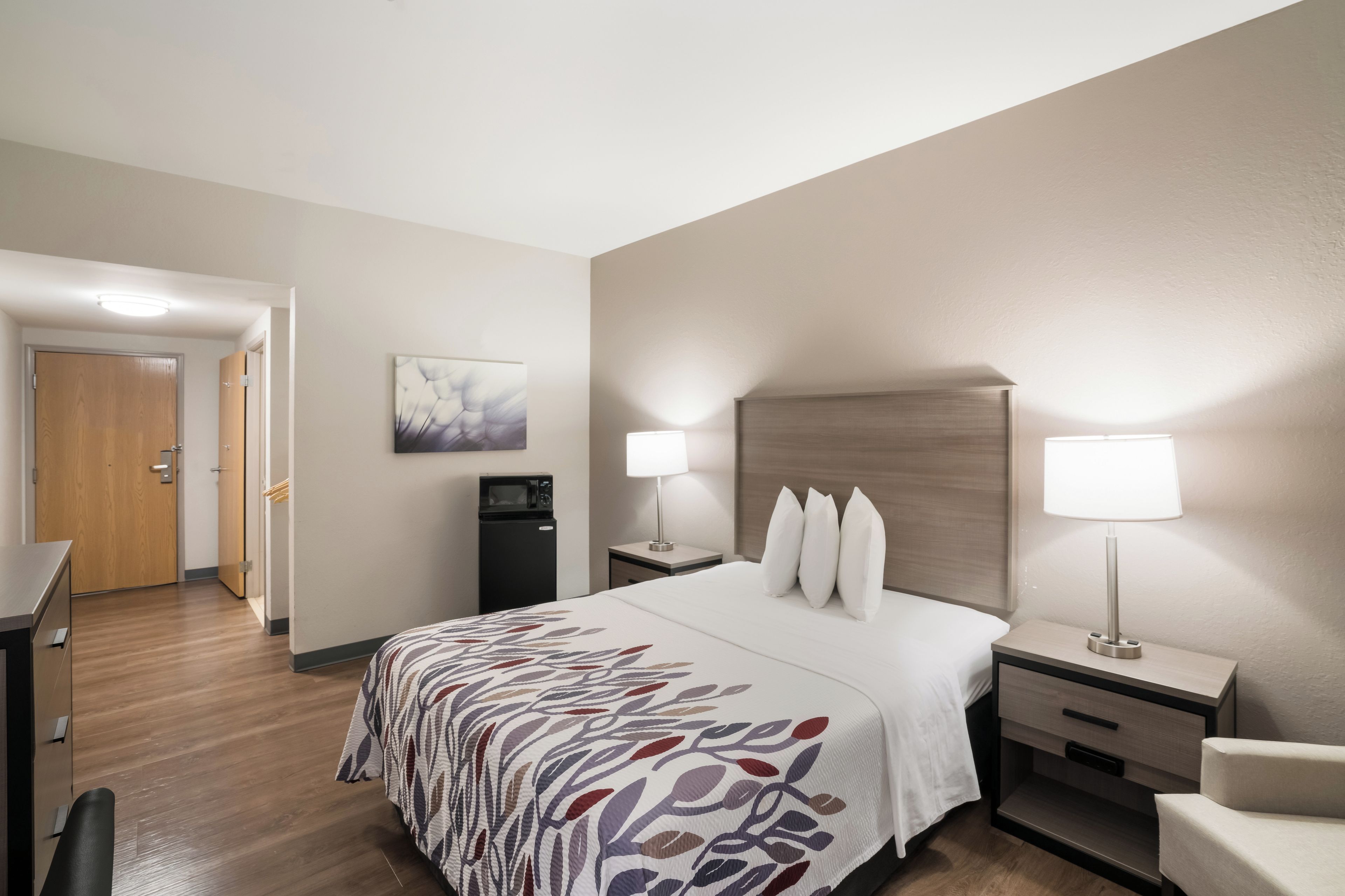 Habitación de hotel con cama, mesitas de noche, escritorio, silla, microondas, refrigerador y puerta.