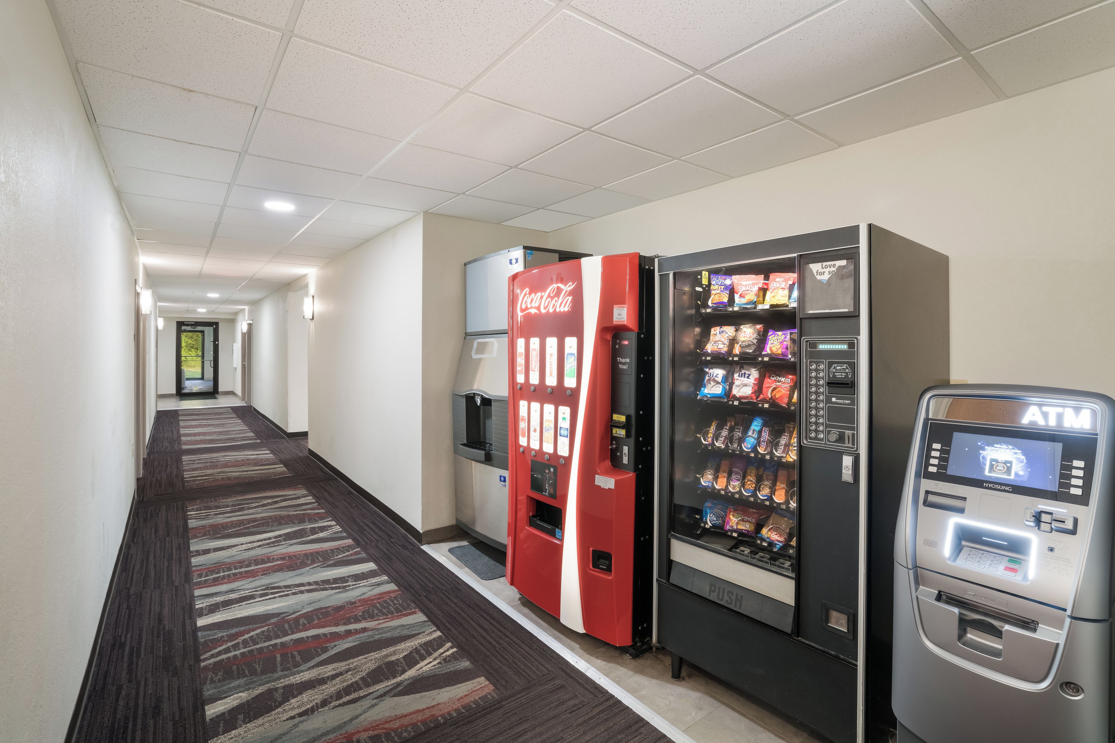 Pasillo de hotel con máquinas expendedoras, incluyendo un refrigerador de Coca-Cola, una máquina de aperitivos y un cajero automático.
