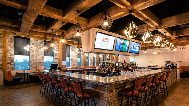 A bar area with a long counter, multiple bar stools, hanging light fixtures, and a screen displaying content. - Deerfoot Inn & Casino (Calgary)