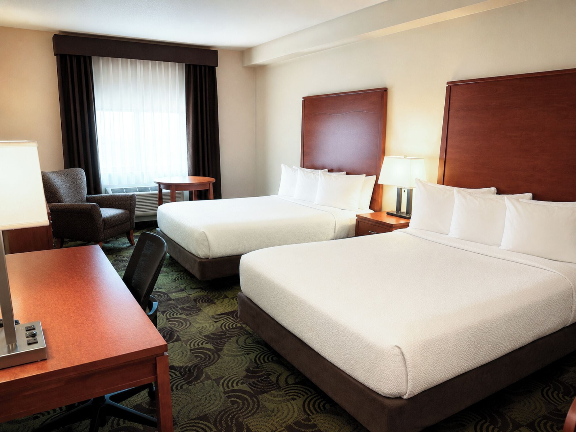 A hotel room with two beds, a desk, a chair, a small table, and a window with curtains.