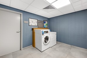 A laundry room with two front-loading washers and dryers, a storage area, and a wall-mounted sign with instructions. - Homewood Suites by Hilton Laredo at Mall del Norte (Laredo)