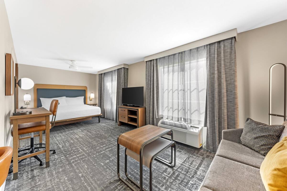 Ett hotellrum med en säng, ett skrivbord, en stol, en TV, en soffa och stora fönster med gardiner.
