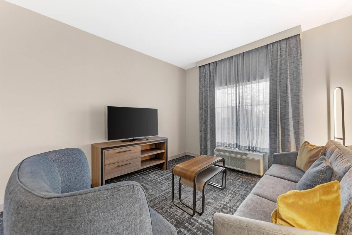 Ett modernt hotellrum med en soffa, en TV, ett litet bord och ett fönster med gardiner.