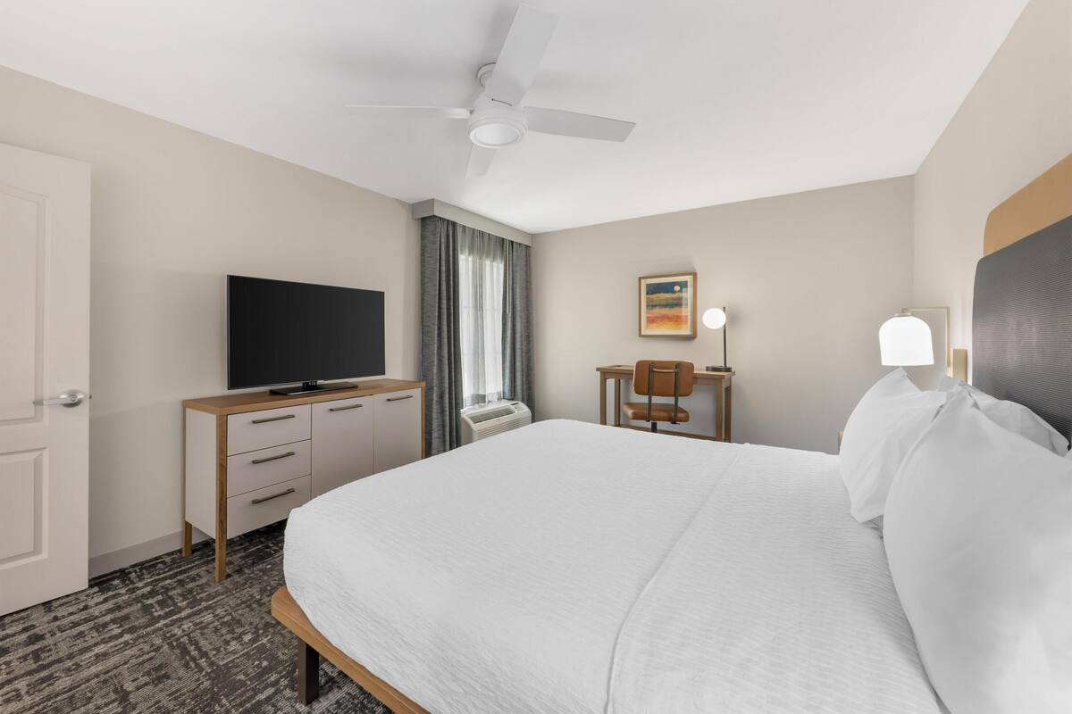 Ett hotellrum med en säng, en tv på en byrå i trä, ett skrivbord med en stol, en takfläkt och ett fönster med gardiner.