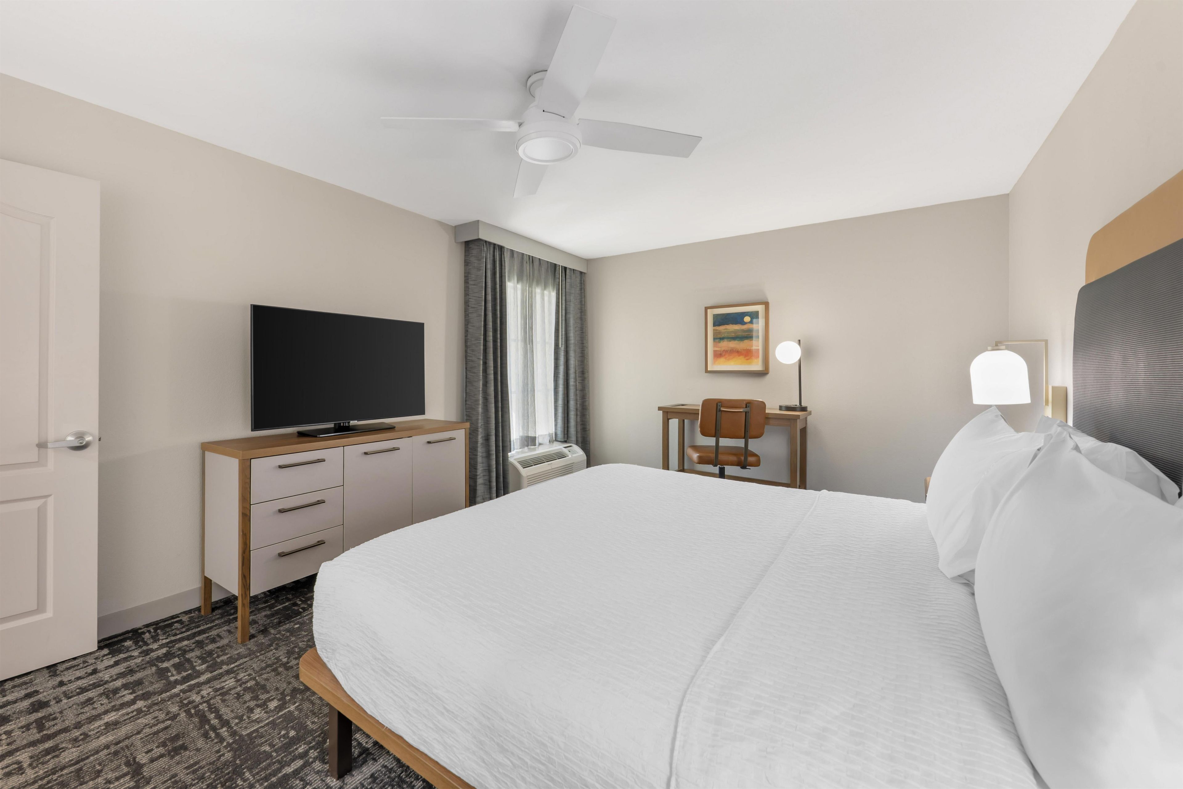 Habitación de hotel con una cama, un televisor sobre una cómoda de madera, un escritorio con una silla, un ventilador de techo y un cuadro en la pared.