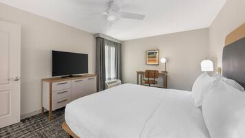 Habitación de hotel con una cama, un televisor sobre una cómoda de madera, un escritorio con una silla, un ventilador de techo y un cuadro en la pared.