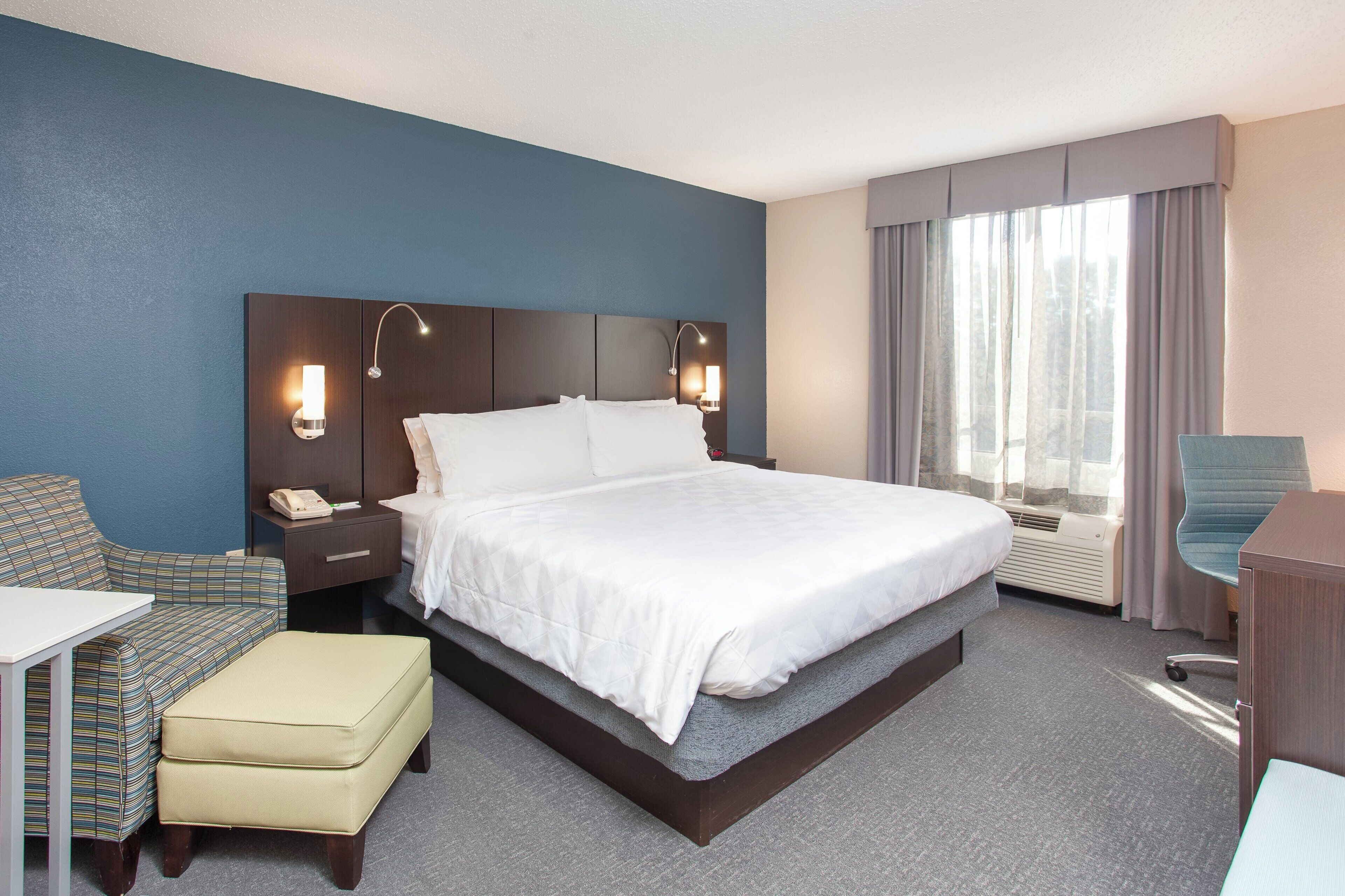 A hotel room with a bed, bedside table, lamp, chair, and a desk.