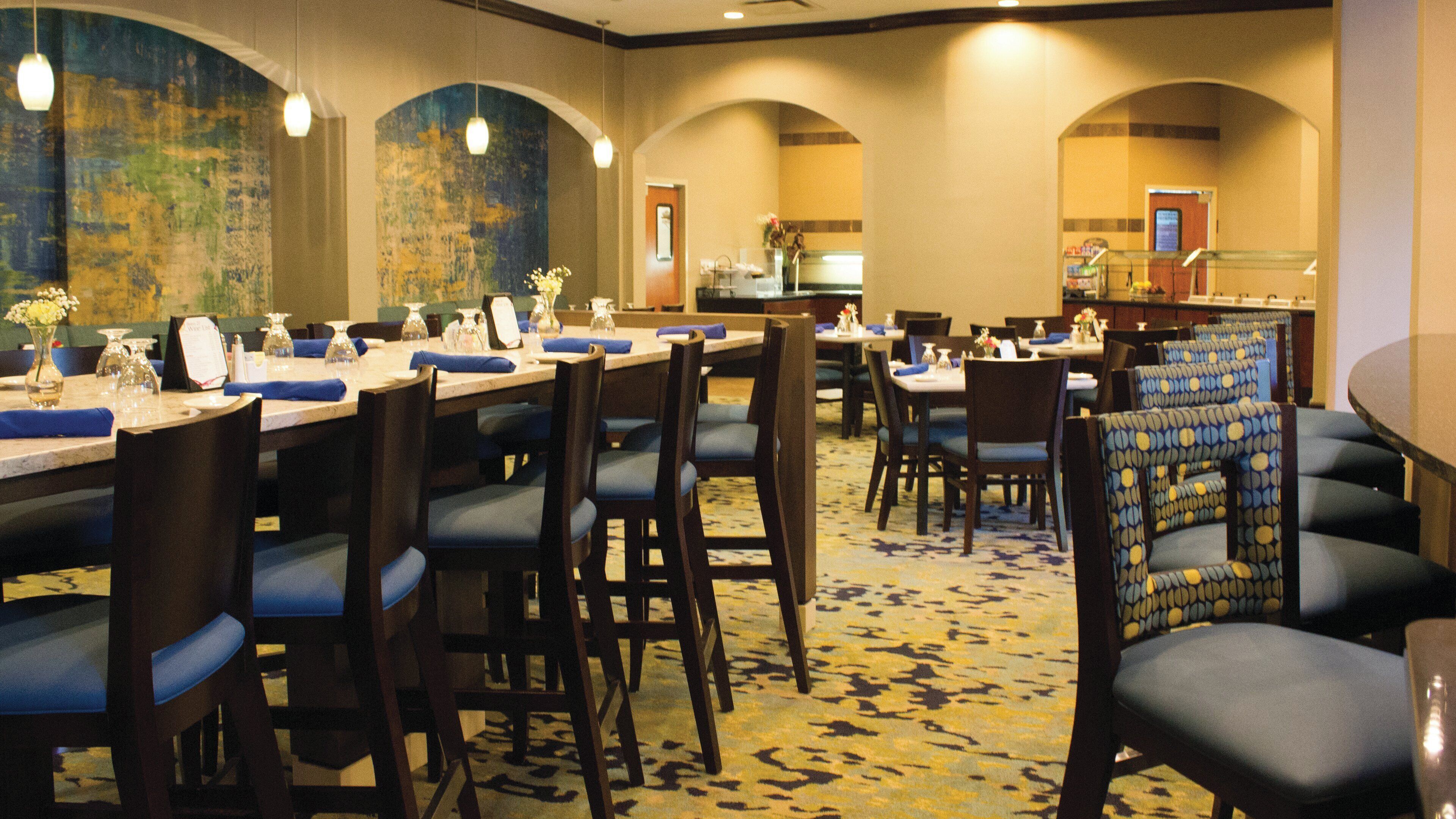 A hotel dining area with multiple tables and chairs, a patterned carpet, and a large mural on the wall.