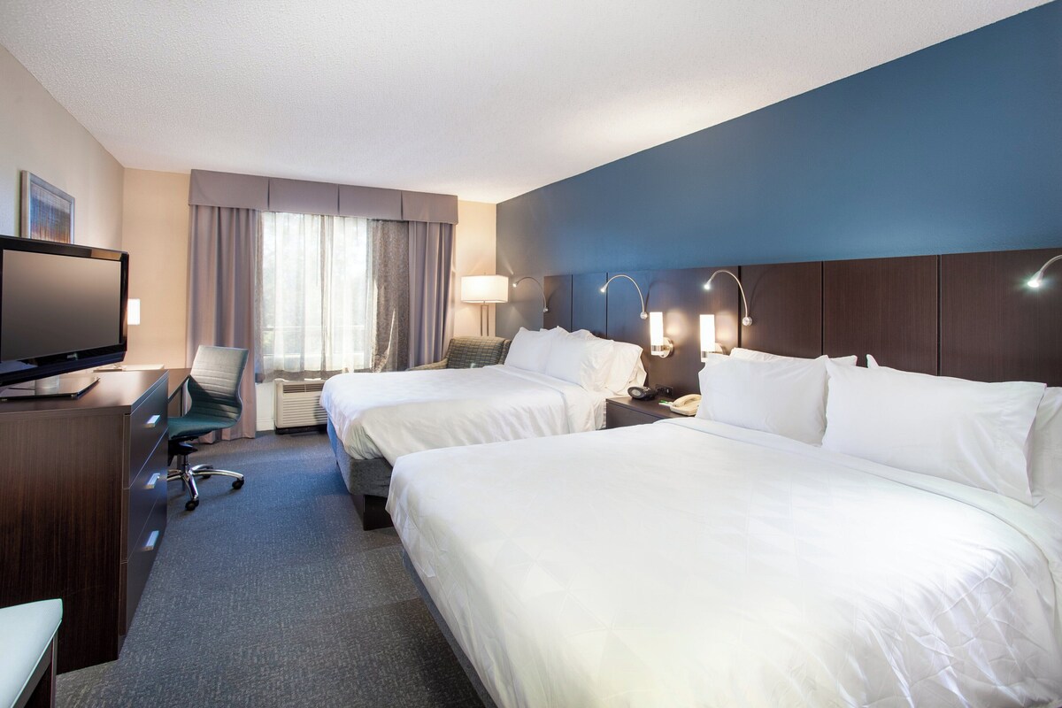 A hotel room with two beds, a desk, a TV, and a window with curtains.