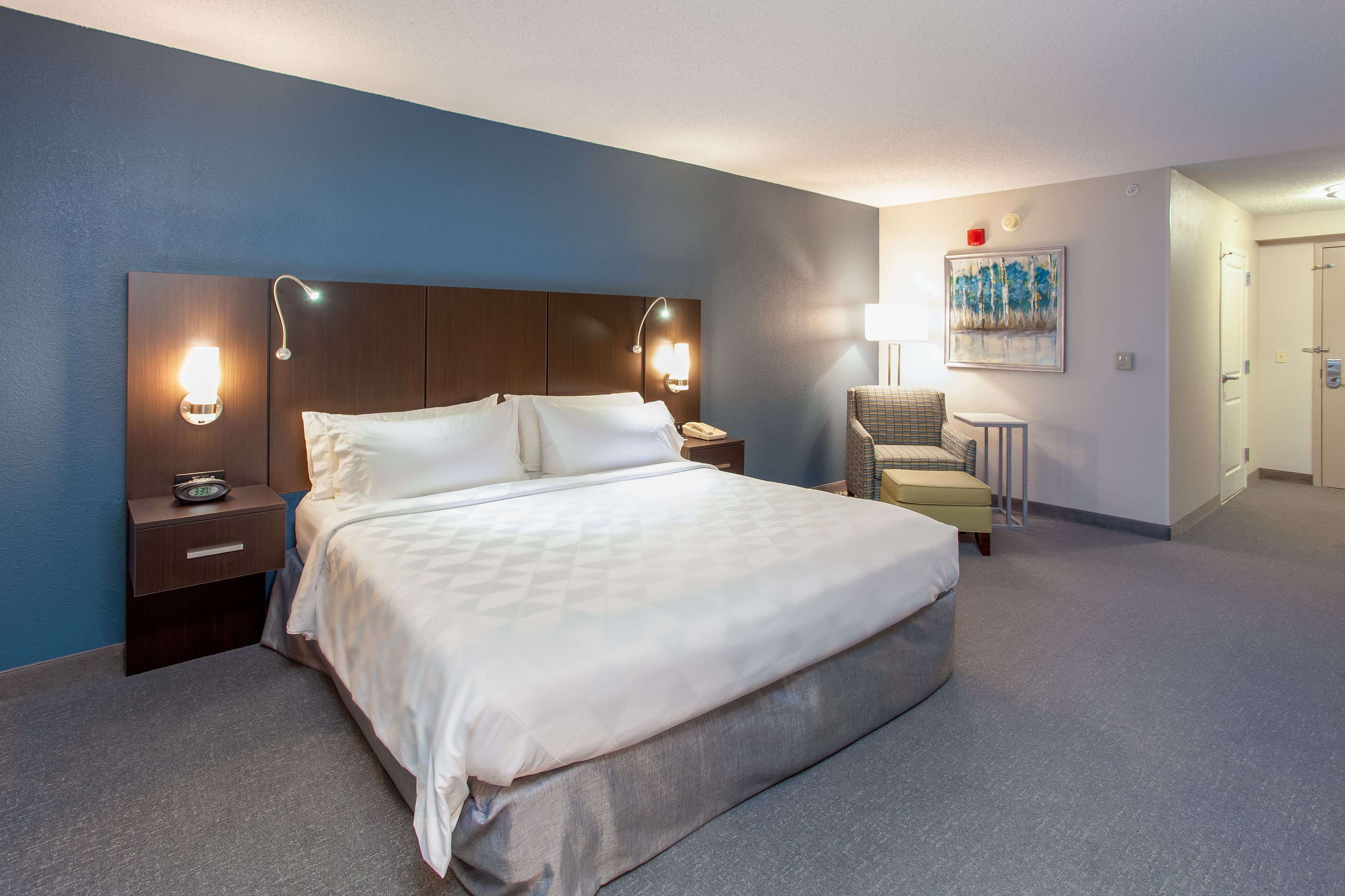 Una habitación de hotel con una cama grande, dos mesitas de noche con lámparas, una silla y un cuadro en la pared.