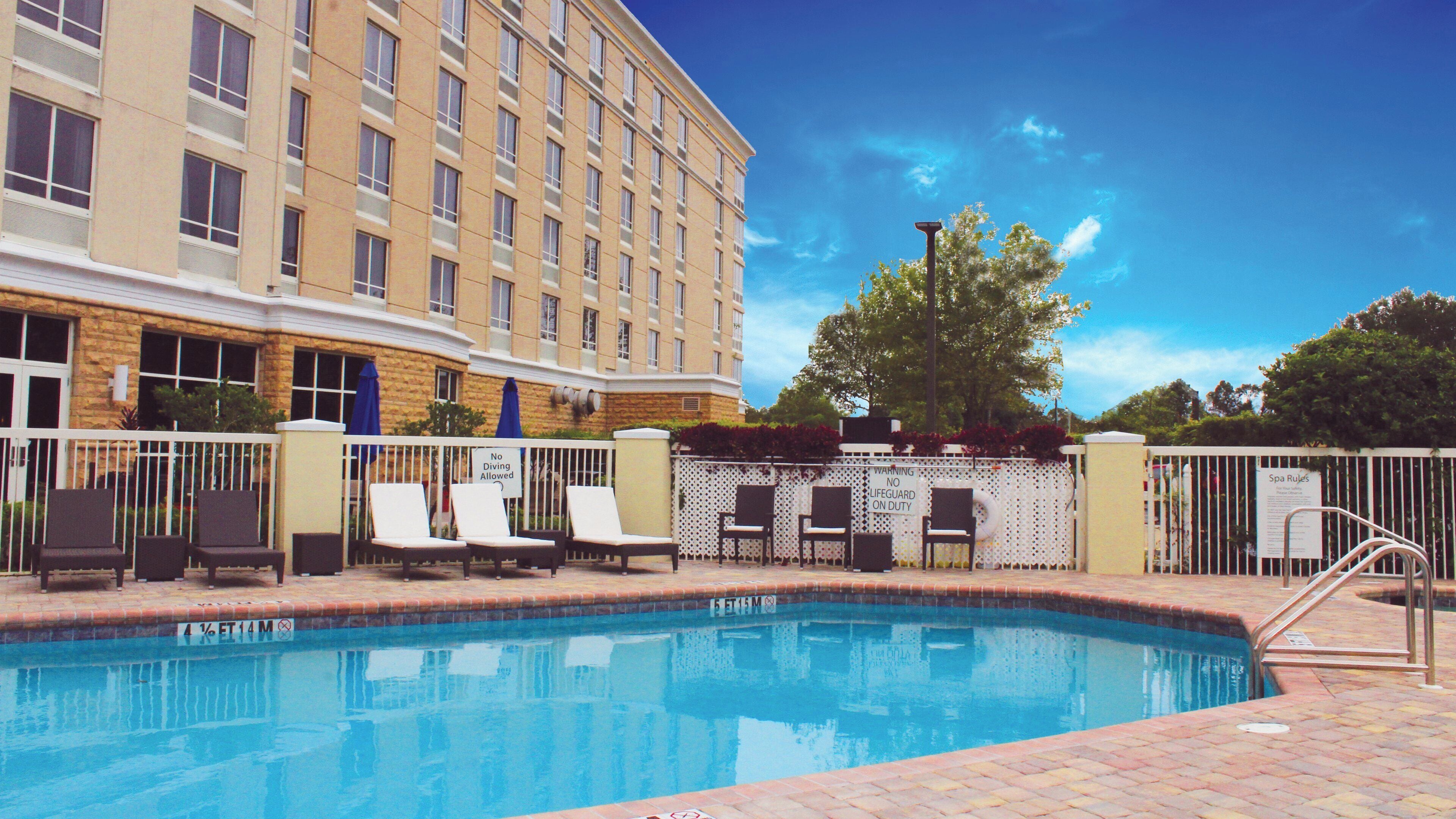 A hotel pool area with lounge chairs, a fence, and a building in the background.