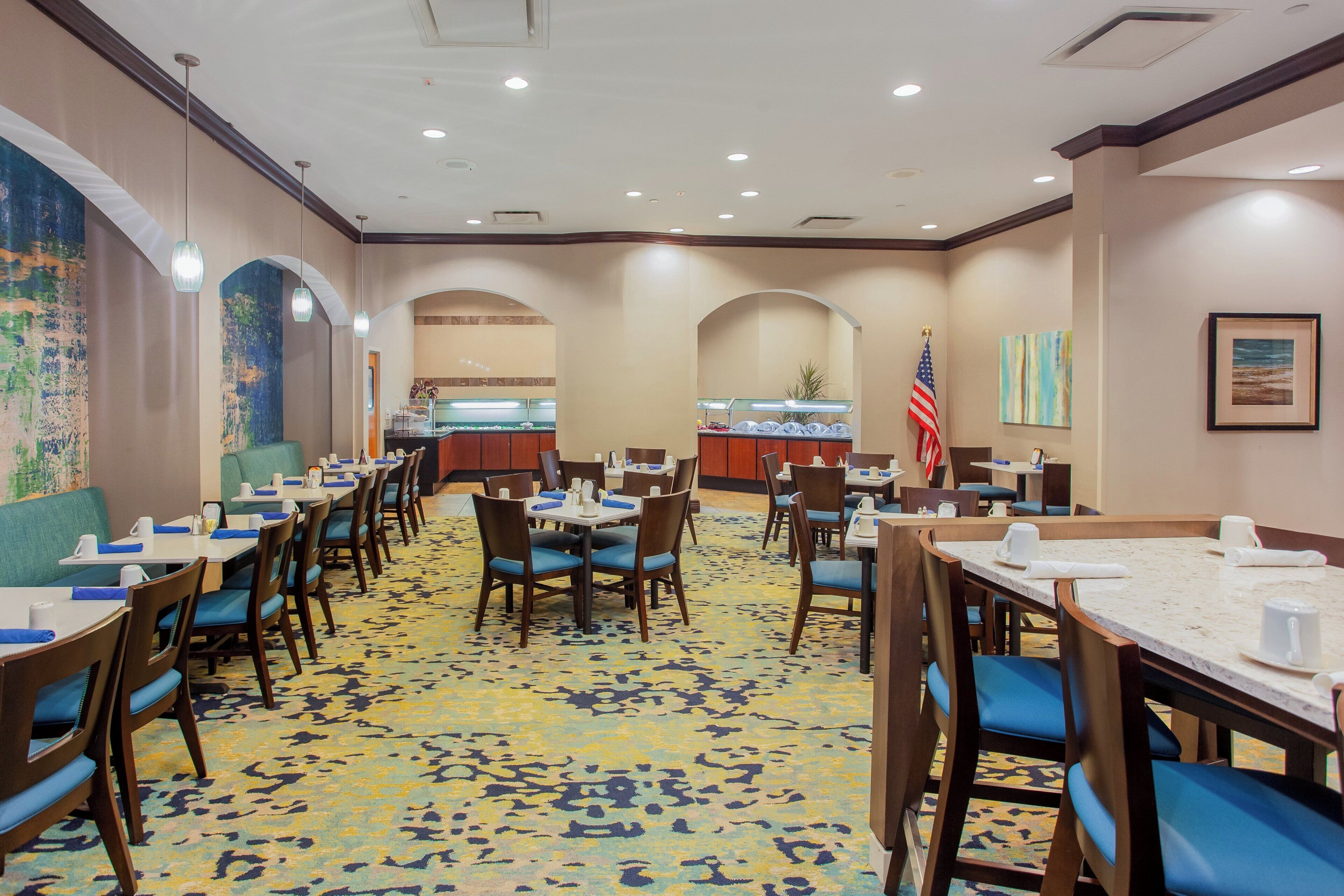 Une salle à manger d’hôtel avec plusieurs tables et chaises, une moquette à motifs et un drapeau américain.