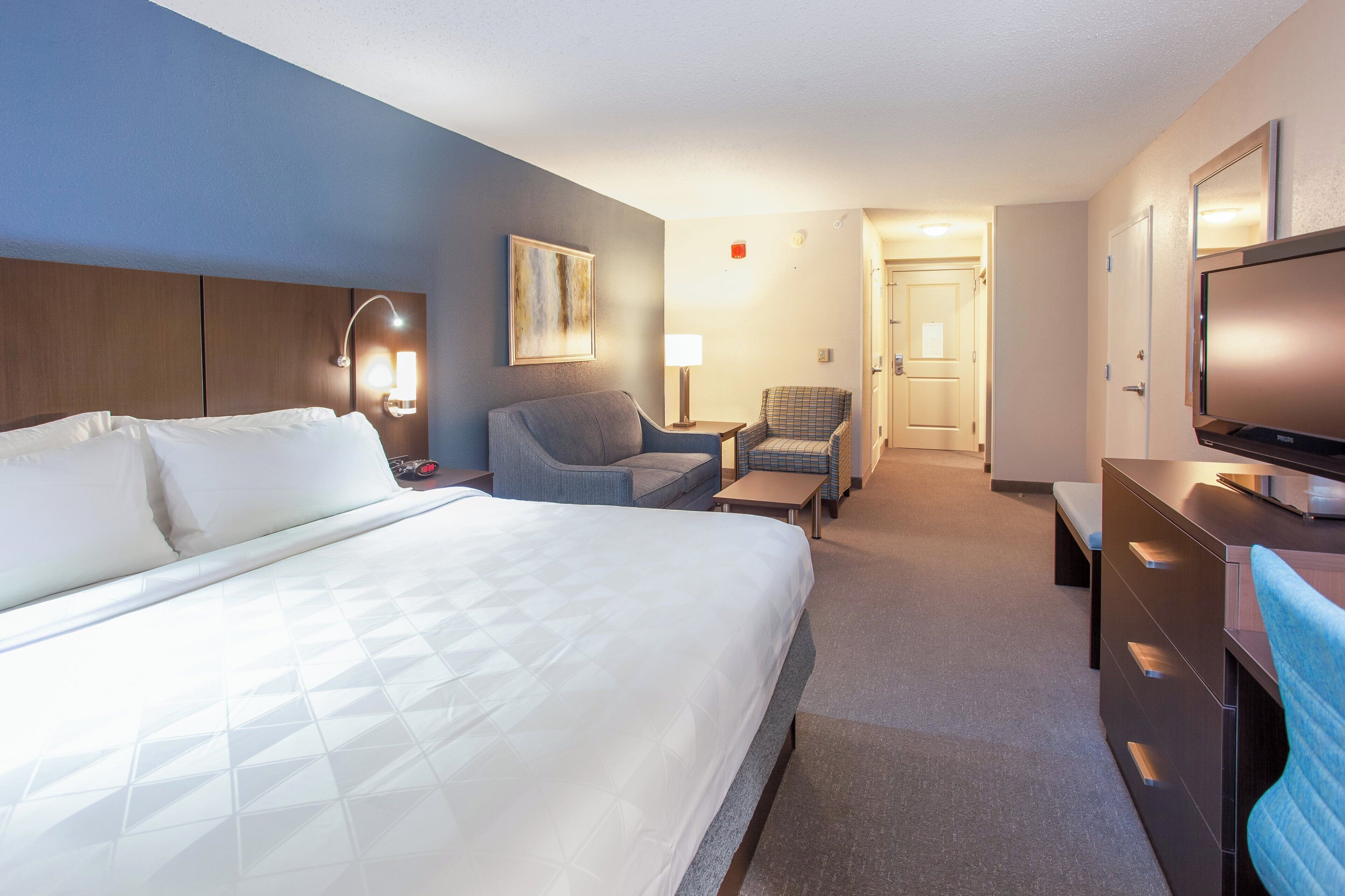 A hotel room with a large bed, a desk, a chair, a television, and a door leading to another room.