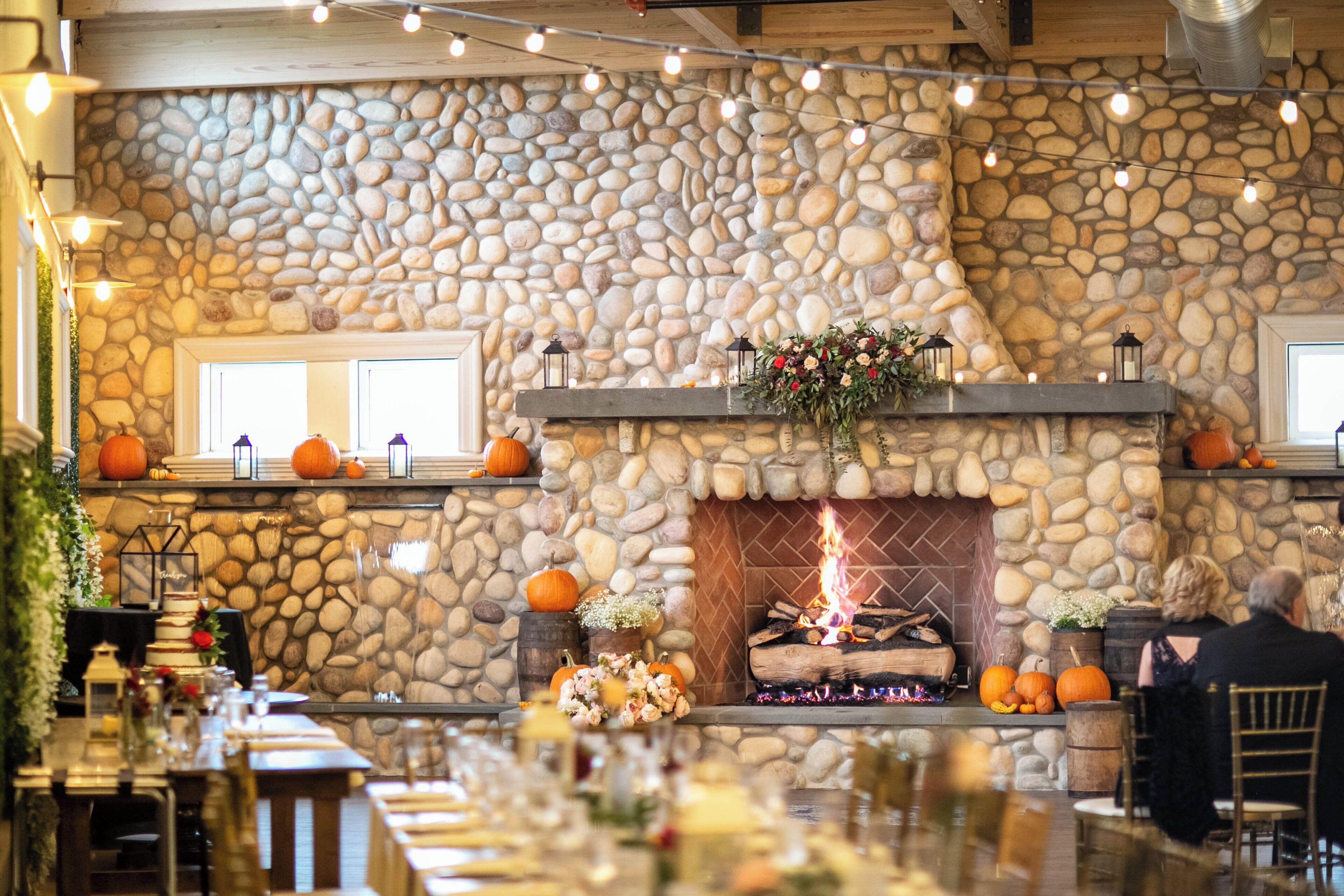 A stone fireplace with a fire, surrounded by pumpkins and flowers.
