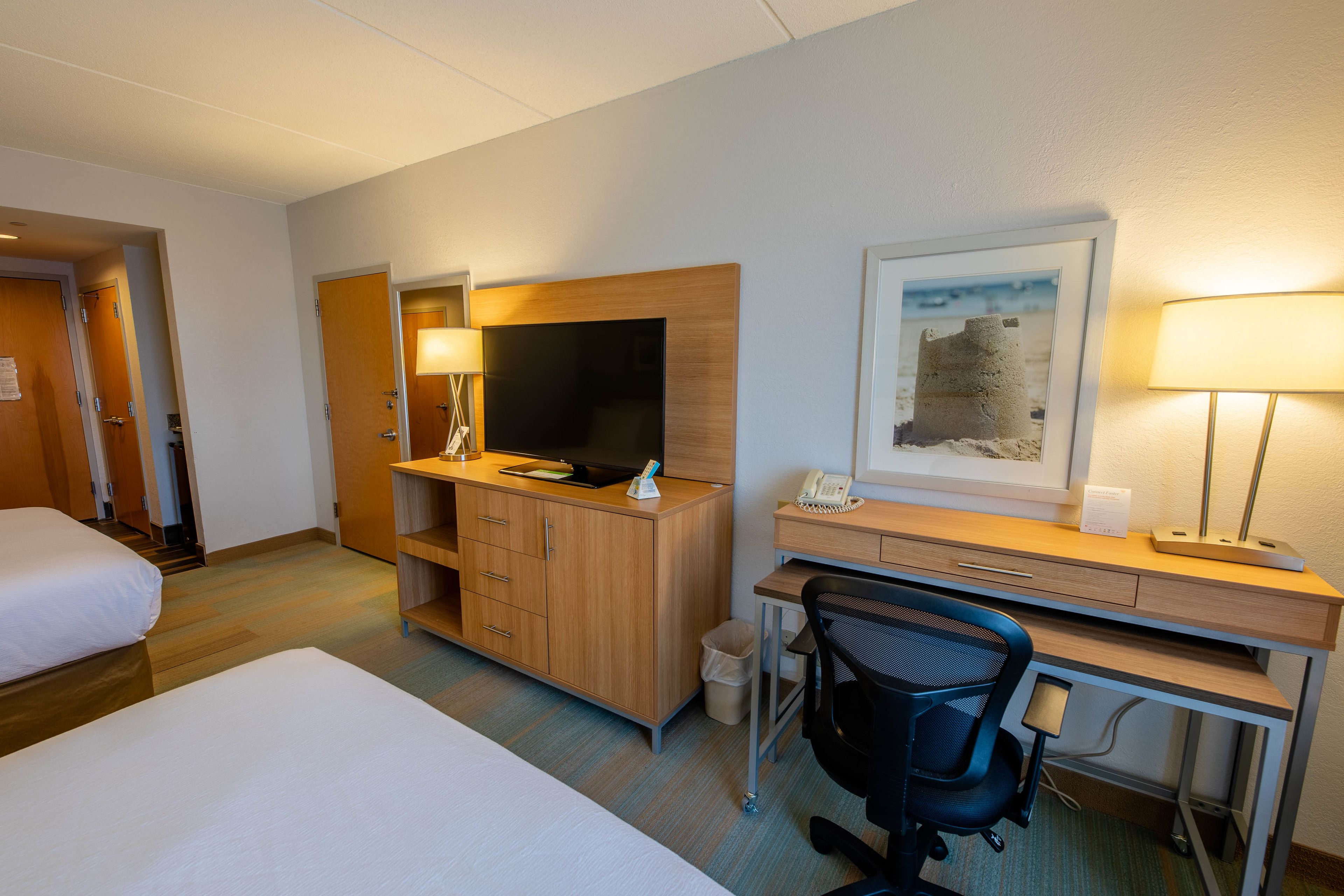Habitación de hotel con cama, escritorio con computadora, silla, televisor y un cuadro en la pared.