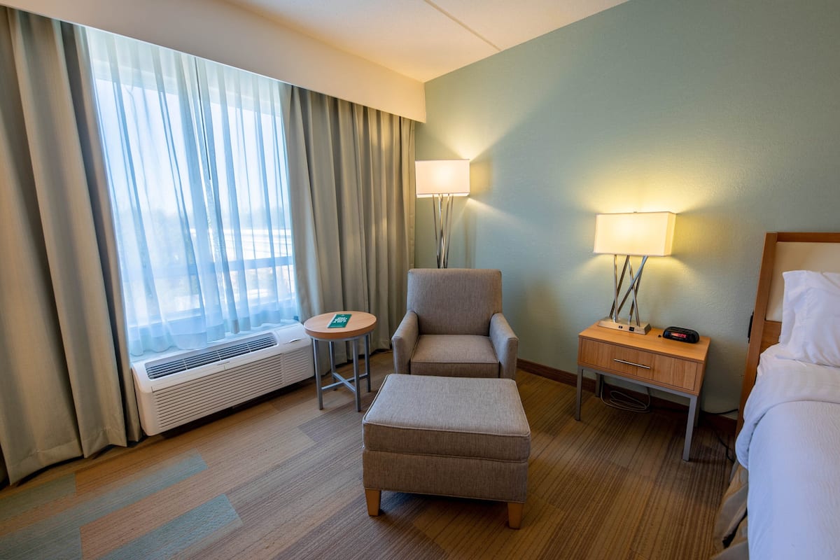A hotel room with a bed, an ottoman, a chair, a nightstand with a lamp, a heater, and a window with curtains.