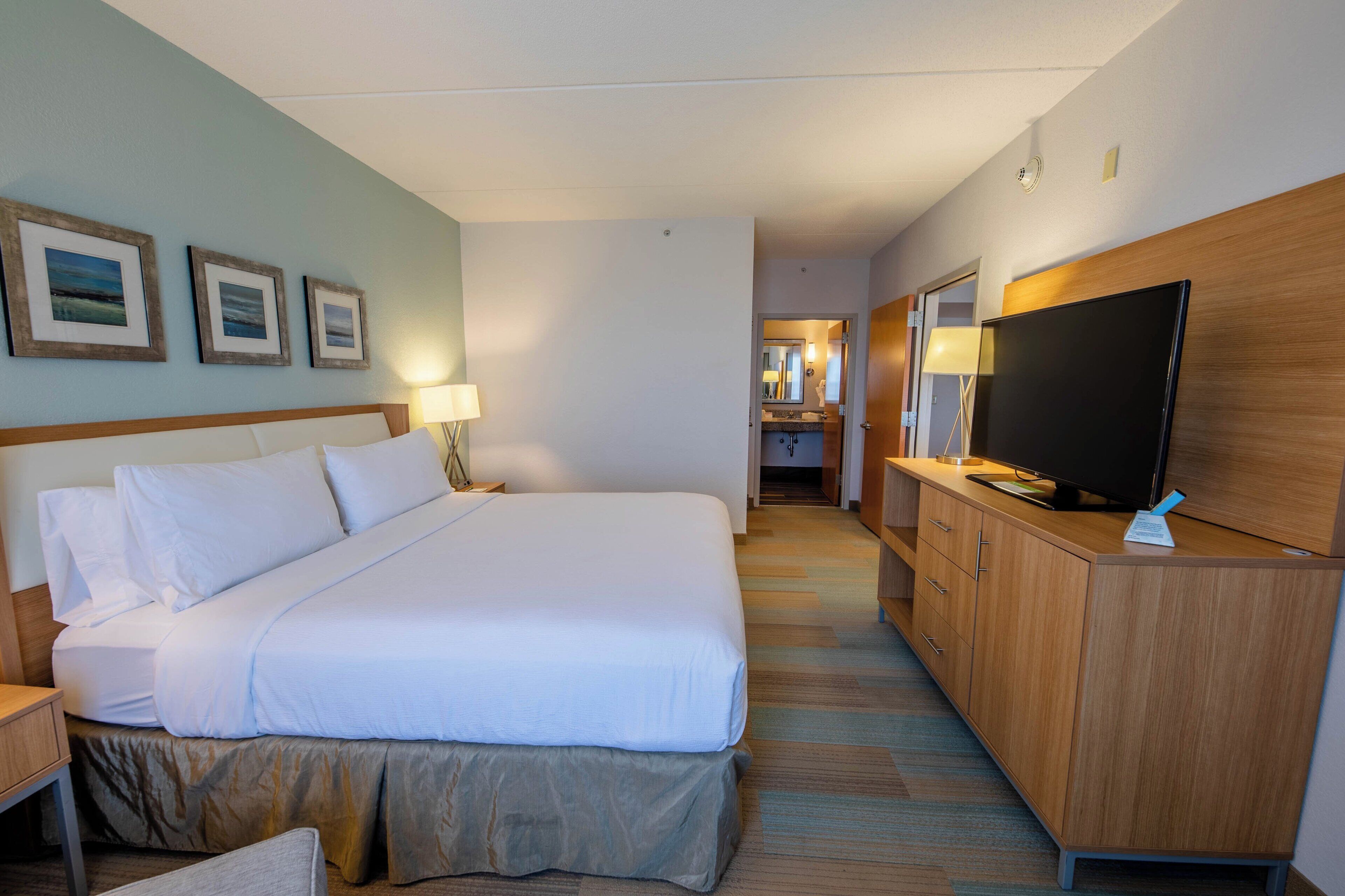 Habitación de hotel con una cama grande, un televisor sobre una cómoda de madera, un escritorio con una lámpara y cuadros en la pared.