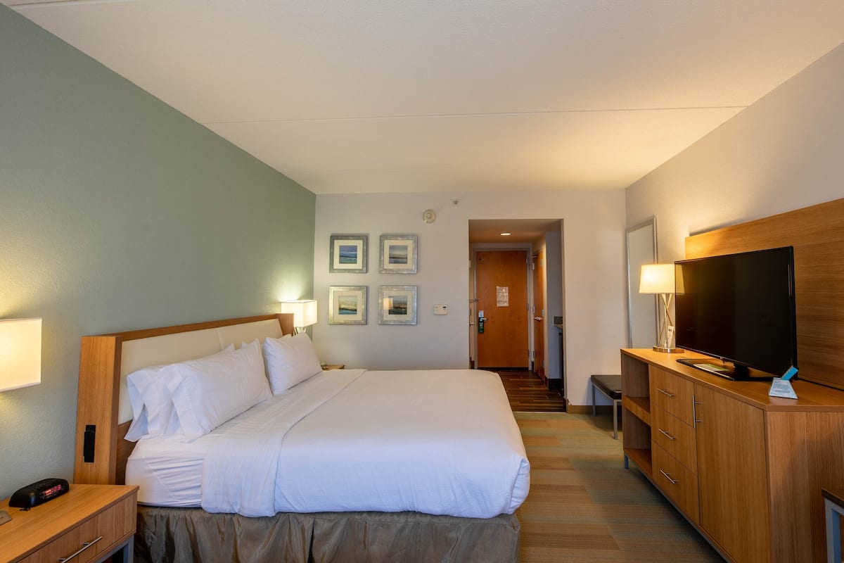 Habitación de hotel con una cama grande, televisor, escritorio y una silla. Hay cuadros en la pared y una puerta que da a otra habitación.