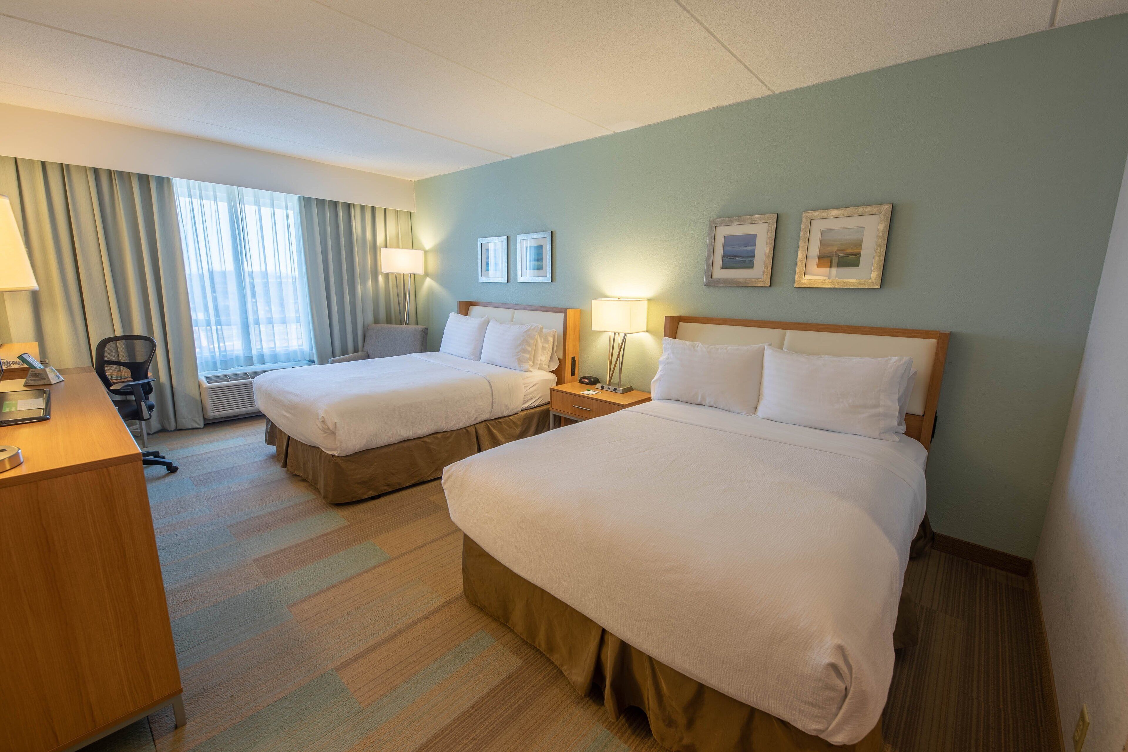 A hotel room with two beds, a desk, and a chair. There are two bedside tables with lamps, a window with curtains, and framed pictures on the wall.
