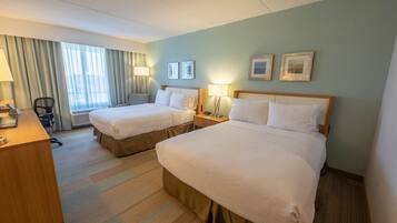 A hotel room with two beds, a desk, and a chair. There are two bedside tables with lamps, a window with curtains, and framed pictures on the wall.