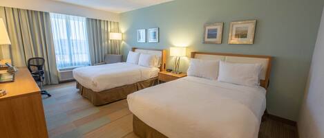Ein Hotelzimmer mit zwei Betten, einem Schreibtisch und einem Sessel. Es gibt zwei Nachttische mit Lampen, ein Fenster mit Vorhängen und gerahmte Bilder an der Wand.