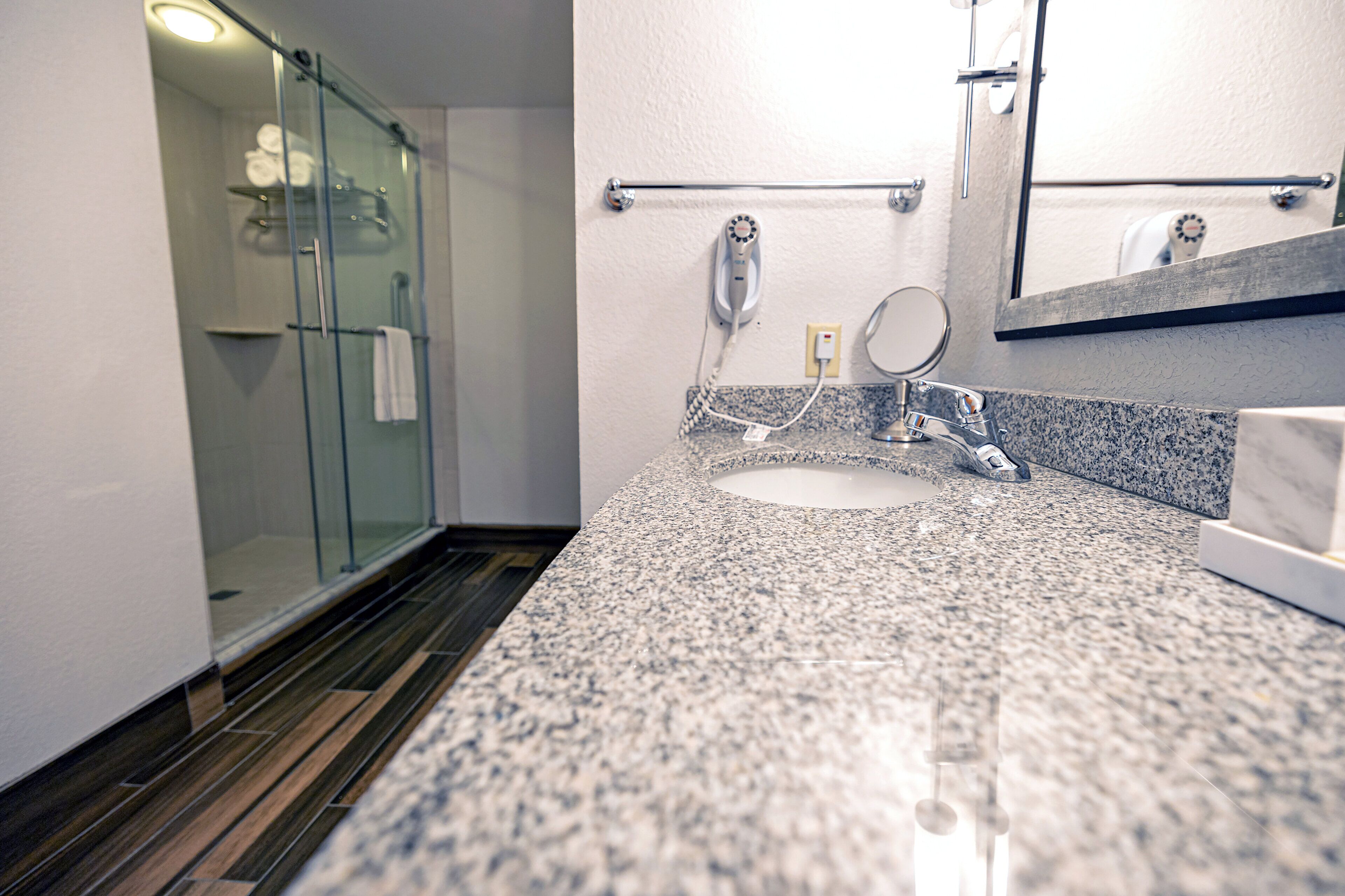 A bathroom with a glass shower enclosure, a granite countertop sink, a handheld mirror, and a towel rack.