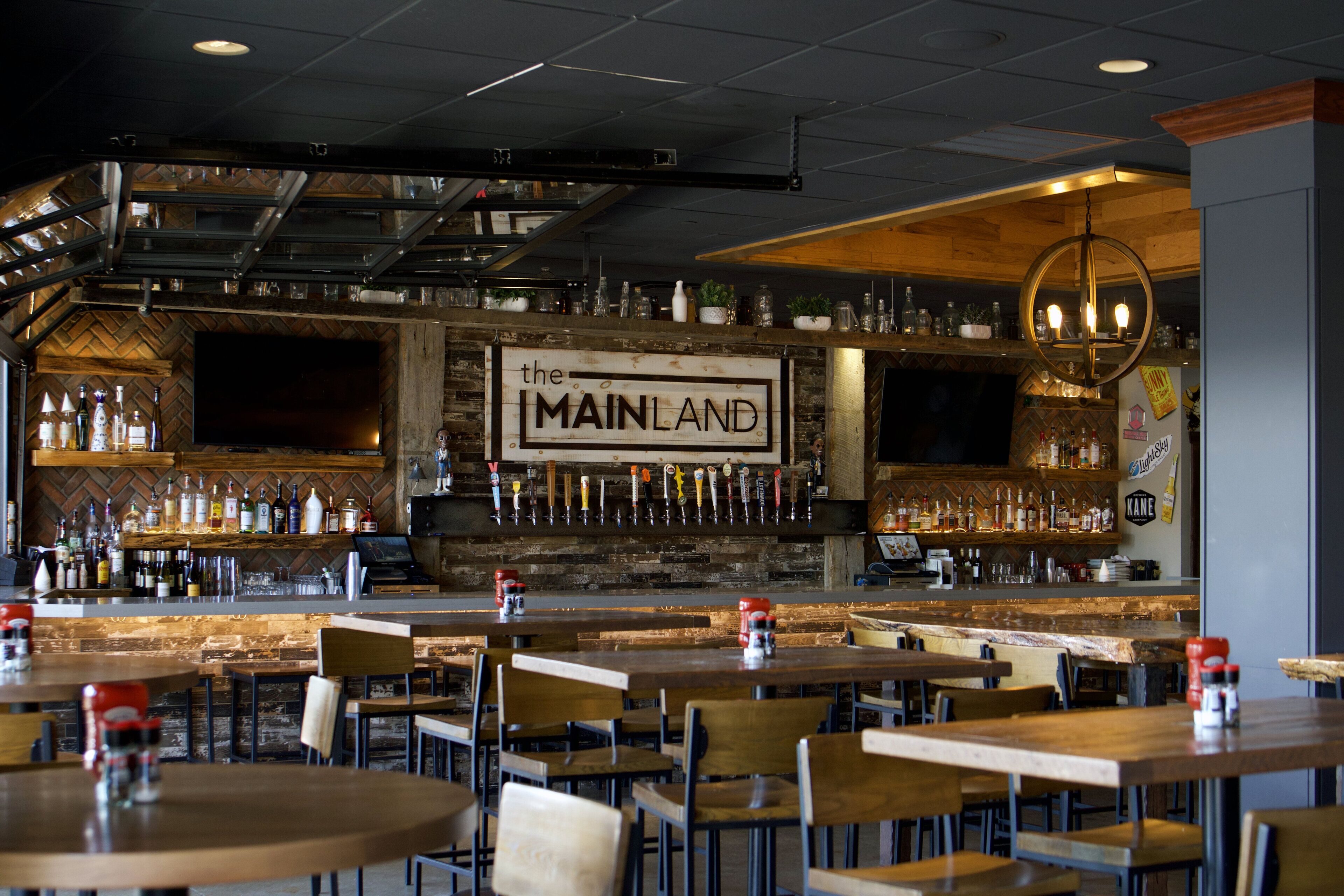Zona de bar con un letrero que dice 'MAINLAND', mesas y sillas de madera y una variedad de botellas en estanterías.