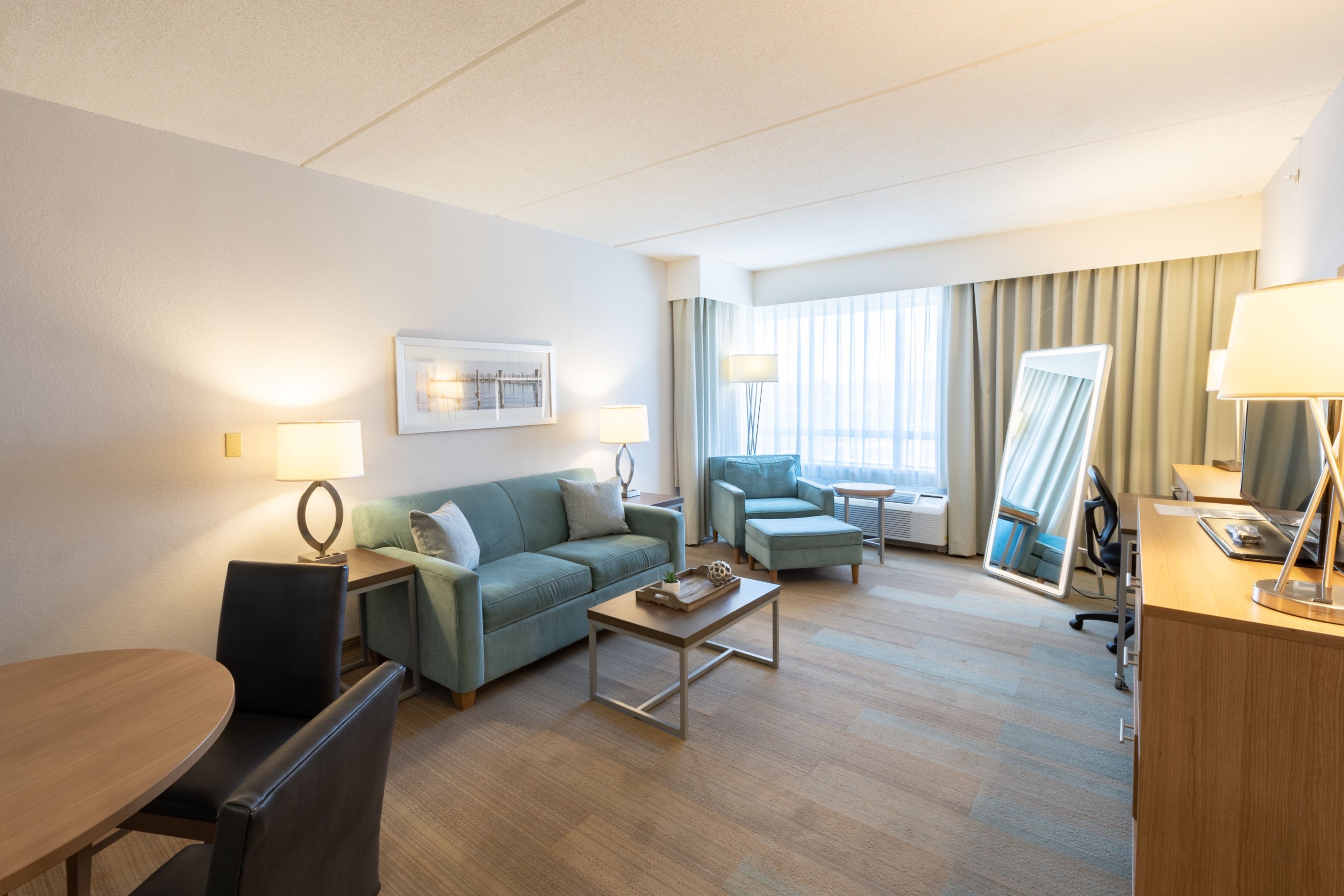 Una habitación de hotel con sofá, sillón, escritorio y un ventanal con cortinas.