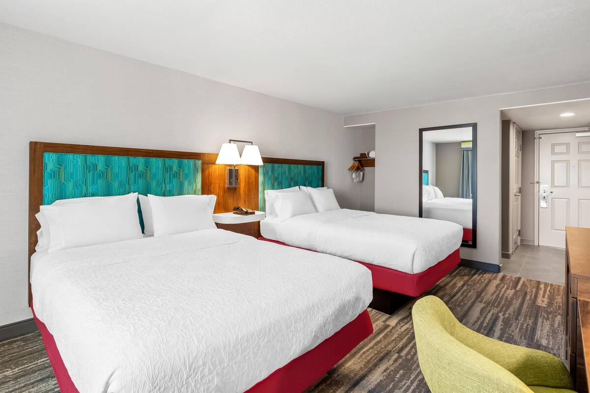 A hotel room with two beds, a nightstand with a lamp, a chair, and a door leading to another room.