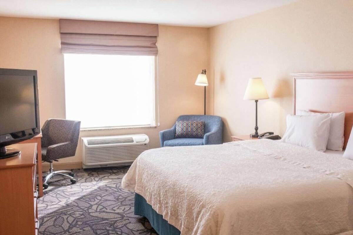 A hotel room with a bed, a desk with a television, a chair, a lamp, and a window with a view.