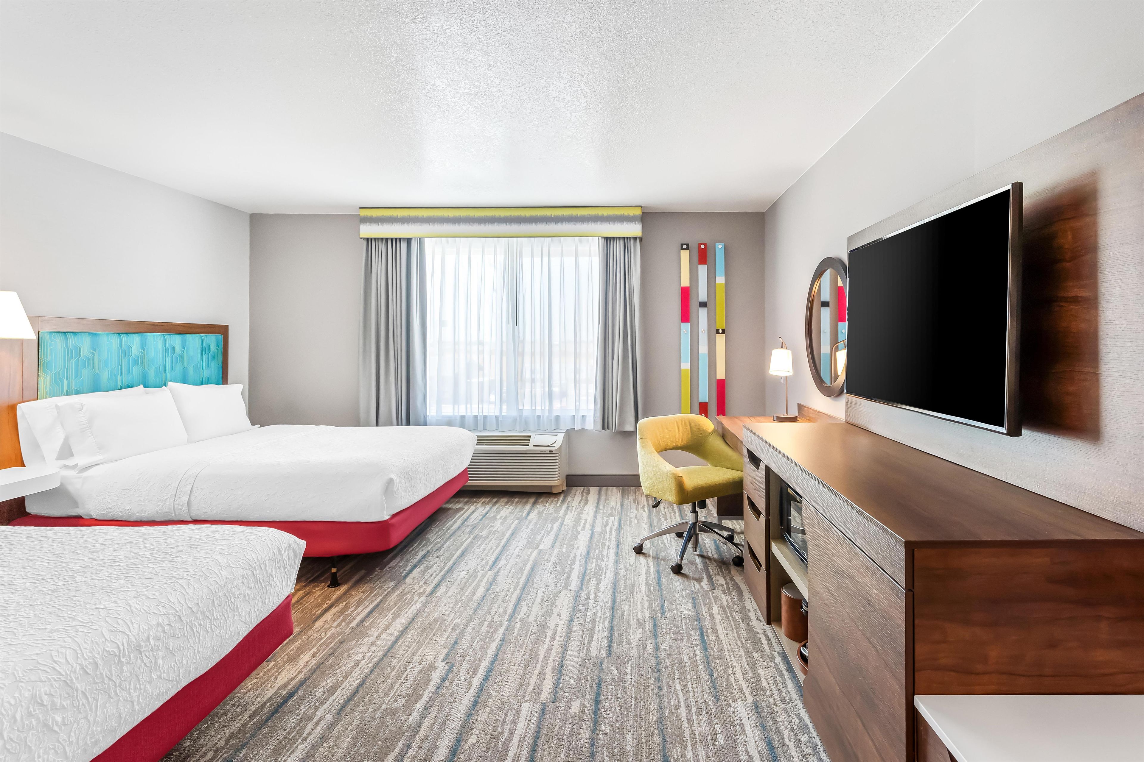A hotel room with two beds, a desk, a chair, a television, and a large window with curtains.