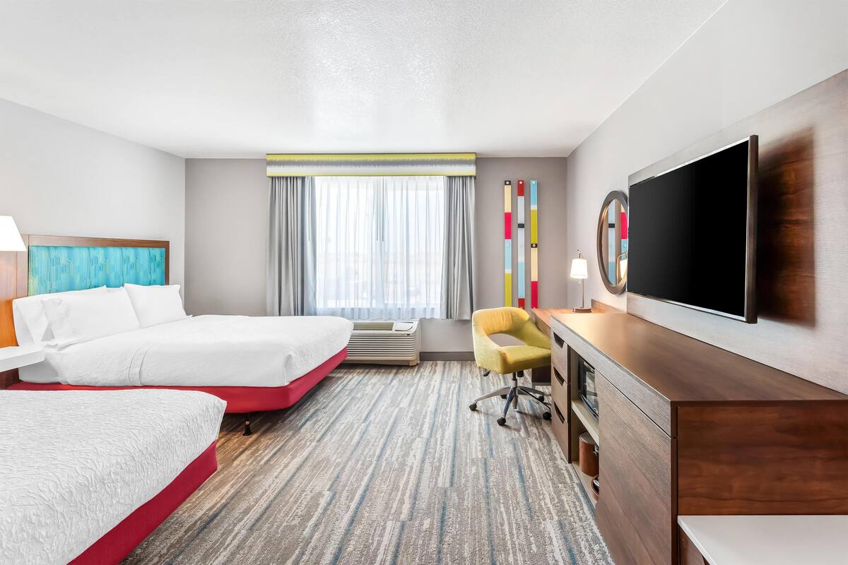 A hotel room with two beds, a desk, a TV, and a large window with curtains.
